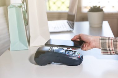 Kadın telefonla ödeme yapıyor. NFC ödeme terminali beyaz bir masada. Kredi kartı ya da telefon faturası artı bankacılık aygıtı, kart makinesi.