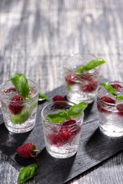 Siyah taş tepside ahududu ve fesleğenli dut kokteyli. Ahşap masada bir bardak maden suyu.