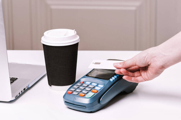 Woman pay by card. NFC payment terminal and take away cup of coffee with chocolate muffin on a white table. Credit card or phone pay pos banking device, card machine.