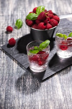 Siyah taş tepside ahududu ve fesleğenli dut kokteyli. Ahşap masada bir bardak maden suyu.