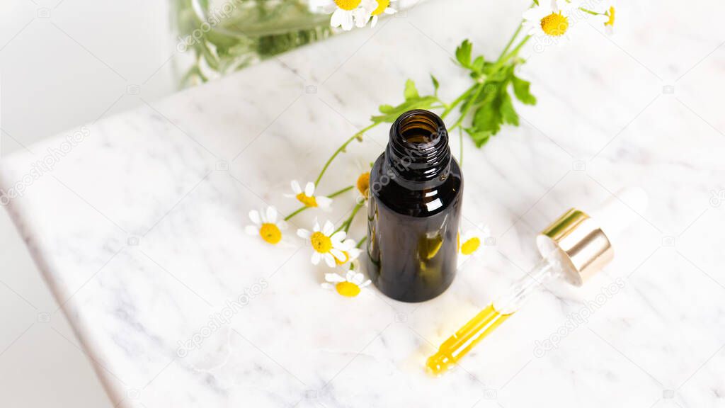 Glass brown bottle of essence oil or serum and with chamomile flowers on marble board. Moisturizing face cream and camomile on white background