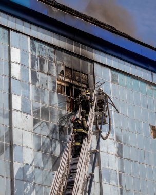 Rusya, Kaluga - SEPTEMBER 08, 2020: Koruyucu giysili ve miğferli bir itfaiye ekibi merdivenlerde, kırık pencereli ve mavi çatılı bir binadaki yangını söndürüyorlar..