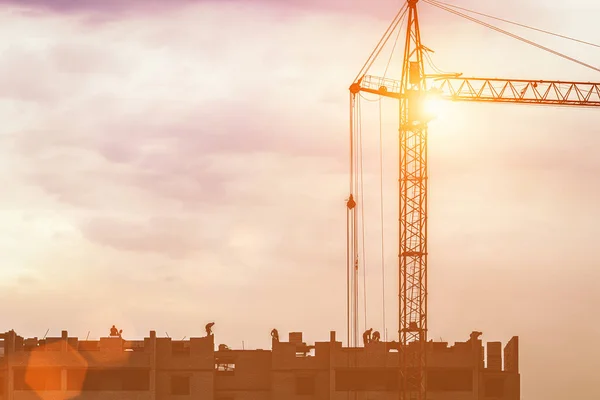 Silhouette of a team of construction workers in and a crane constructing a building on the background of the evening sunset sky.