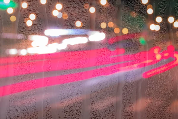 Defocused headlights from moving cars behind rainy, wet glass at night. Abstract concept of rainy autumn weather.