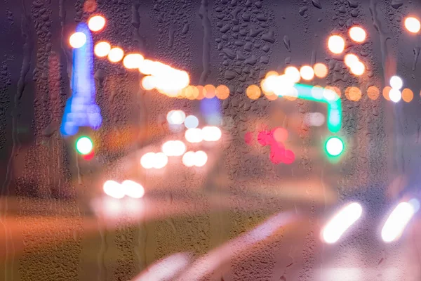 Defocused headlights from moving cars behind rainy, wet glass at night. Abstract concept of rainy autumn weather.