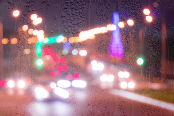 Defocused headlights from moving cars behind rainy, wet glass at night. Abstract concept of rainy autumn weather.