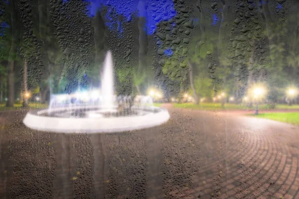Defocused fountain in a night park with lights behind rainy, wet glass at night. Abstract concept of rainy autumn weather.