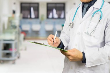 Hastane koridorunda dosyaları ve steteskopu olan erkek doktor elinde pano, reçete yazıyor.