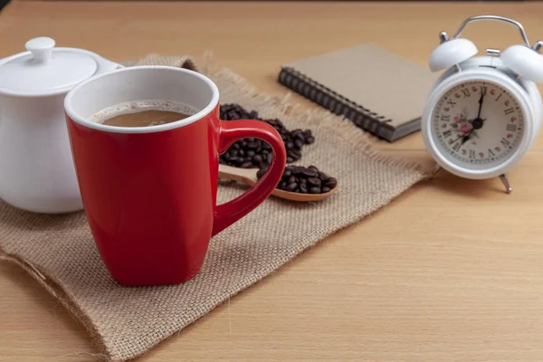 Red coffee cup with coffee beans and white alarm clock on business table.