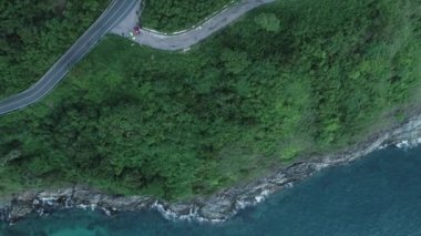 İHA 'nın yukarıdan aşağıya uçuş videosu asfalt yolda akşam vakti tropikal deniz kıyısında dönüyor.