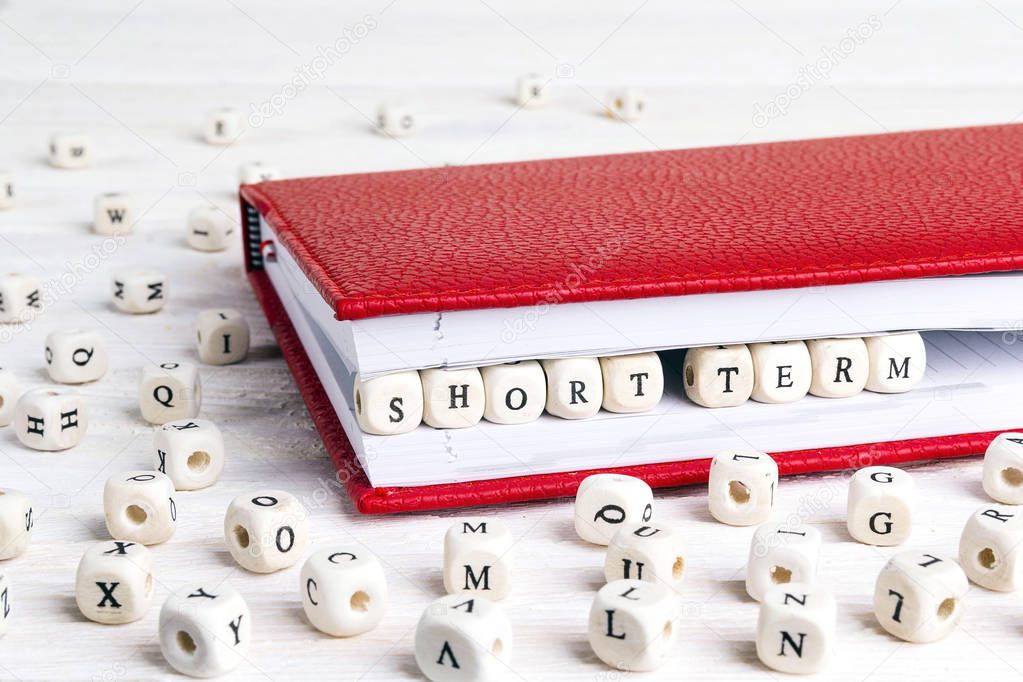 Phrase Short term written in wooden blocks in red notebook on white wooden table. Wooden abc.