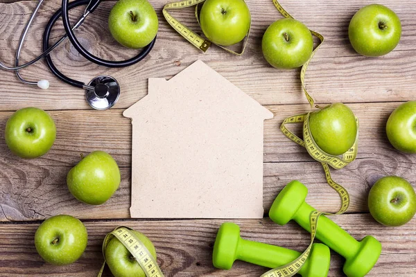 House symbol with green dumbbells and apples with measuring tape and stethoscope on wooden background. Sport, fitness, health and diet concept. Top view.