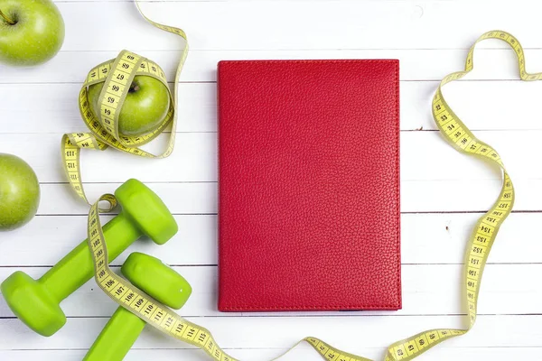 Red notepad with green dumbbells and apples with measuring tape on white wooden background. Sport, fitness and diet concept. Top view.