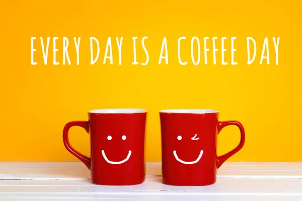 Two red coffee mugs with a smiling faces on a yellow background with the phrase Every day is a coffee day. Happy coffee mugs.