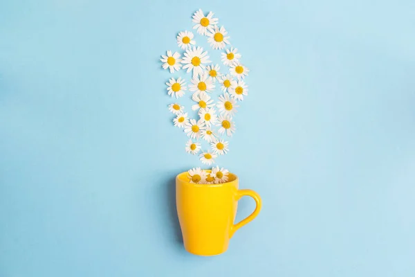 Yellow mug and chamomile flowers on a blue background. Chamomiles come out of the mug like steam. Chamomile tea concept. Flat lay, top view, copy space.