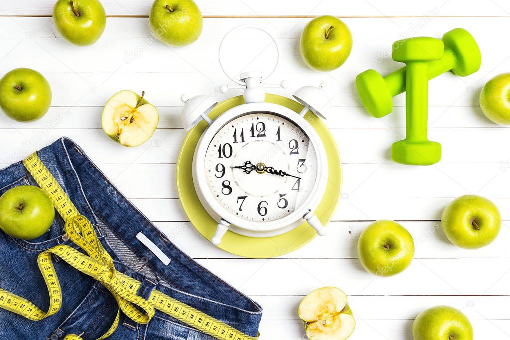 Alarm clock with measure tape, blue womans jeans, green apples and dumbbels on white wooden background. Healthy lifestyle, body slimming and weight loss concept. 