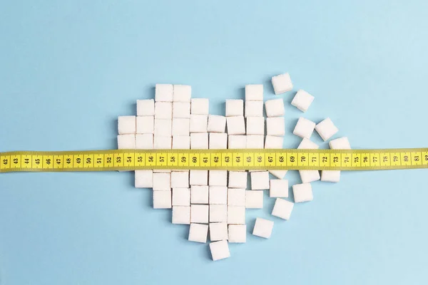 Broken heart made of sugar cubes with measure tape on a blue background.  Diabetes, diet and weight loss concept.