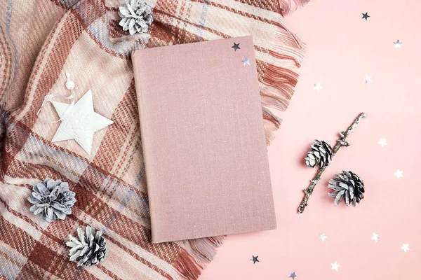 Top-down winter holidays composition with book, plaid, cones and stars on pink background. Copy space for text.