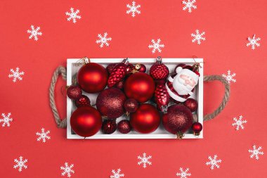 Bir beyaz ahşap tepsi kar taneleri ile kırmızı bir arka plan üzerinde kırmızı Noel oyuncaklar. Üstten görünüm, düz yatıyordu.