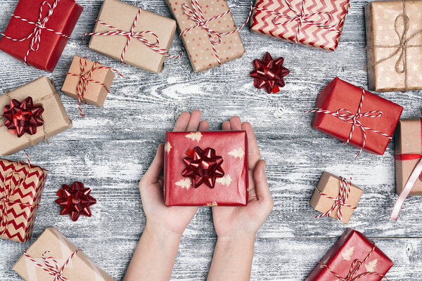 Women's hands with gift box surrounded by wrapped gifts. Top-down Christmas composition on grey wooden background.