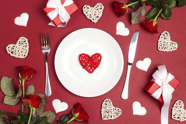 Накрытие праздничного стола со столовыми приборами, цветами роз и подарочными коробками на красном столе. Квартира лежала, сверху вниз композиция. День святого Валентина фон
. 