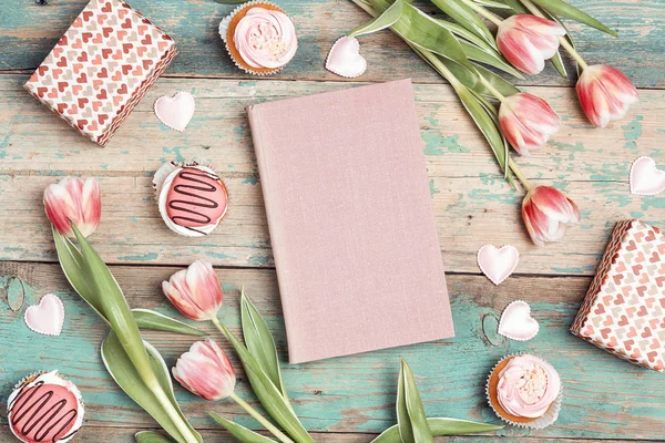 Pink book with tulip flowers, cupcakes and gifts on vintage wooden table with copy space. St. Valentine, Mothers, Womens Day concept. Top down composition.