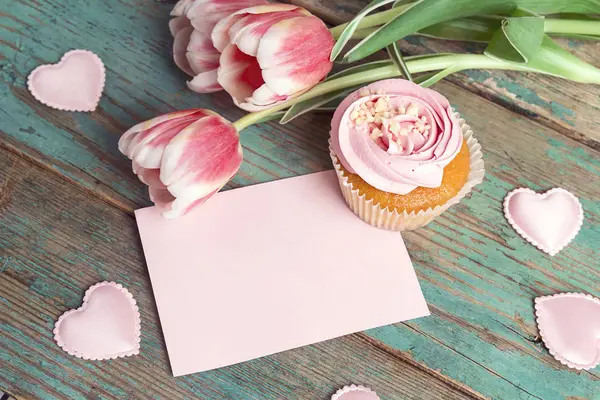 Blank pink card with cupcake, tulips and hearts on turquoise wooden background. Romantic love background. St. Valentine, Mothers, Womens Day concept. Copy space.