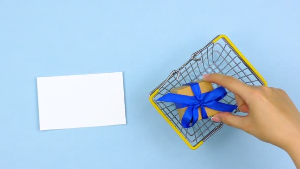女性の手はギフトボックスをショッピングバスケットに入れました 青い背景のテキストのための空白の白い紙のカード — ストック動画