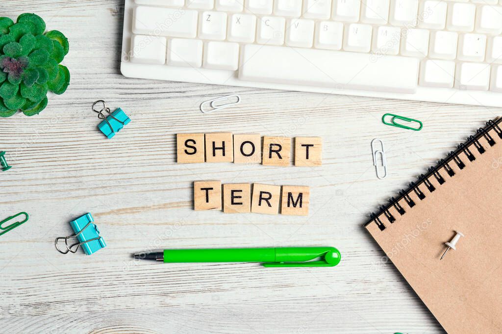 Wooden phrase Short term in workspace  with computer keyboard, supplies  and green plant on light wood background. Top view, flat lay office desk table. Wooden abc.