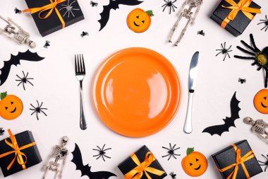 Cadılar Bayramı sofrası portakal tabağı, çatal bıçak takımı, ürkütücü süslemeler ve beyaz masada hediyeler. Üst alt kompozisyon.
