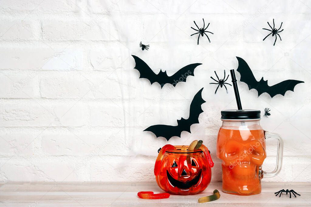 Orange Halloween cocktail in skull glass jars with gummy worm against a white brick wall background. Copy space for text.