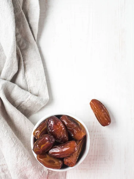 Small bowl of sweet dried dates and linen cloth on white or light gray cement background. Organic dried dates with copy space for text. Top view or flat-lay. Vertical.