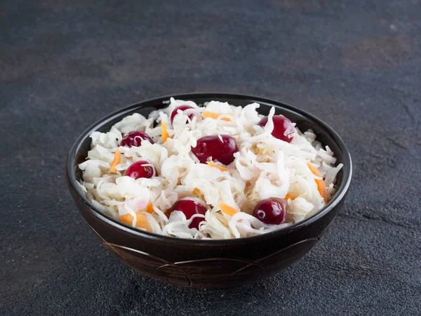 Traditional russian appetizer sauerkraut with cranberry and carrot in dark bowl on black cement background. Fermented cabbage. Russian cuisine and russian kitchen. Copy space.