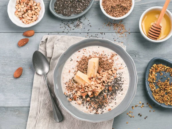 Overnight oats in bowl and ingredients - banana, LSA, chia seeds, almond, honey and pollen on gray wooden table background. Healthy breakfast oatmeal recipe idea. Top view
