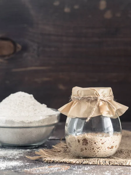 Active rye sourdough starter in glass jar and rye flour on brown wooden background. Starter for sourdough bread. Toned image. Copy space. Vertical