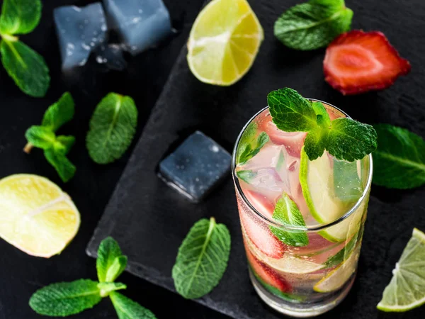 fresh lemonade with strawberry, lime and mint on dark stone background. Cold summer strawberry drink with mint and ice. Strawberry mojito in glass and ingredients