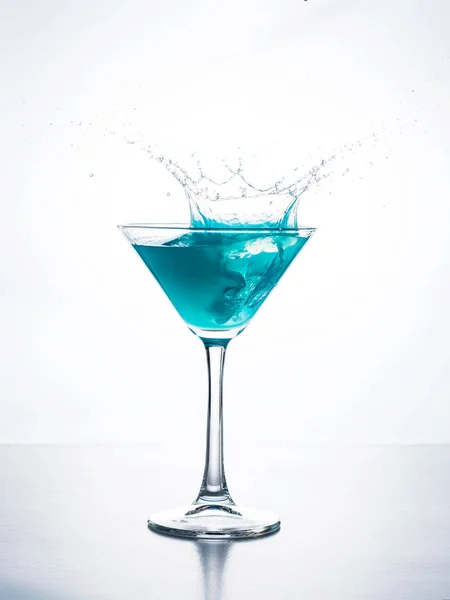 Blue cocktail in martini glass with ice cube splashing into liquid against white background. Blue curacao cocktail with splash on white.