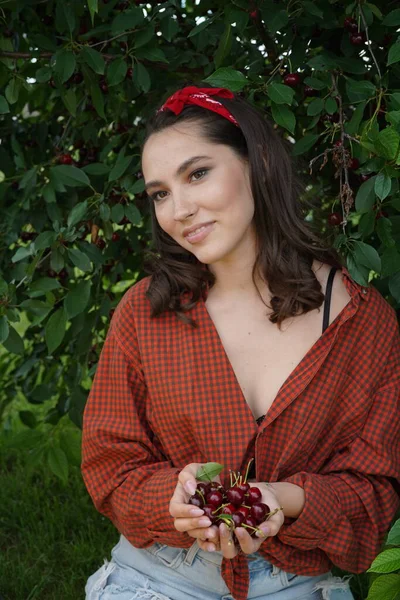 Mulher sorridente com um punhado de cerejas. Jovem morena segurando cerejas frescas e maduras em suas mãos e mostrando-as à câmera . — Fotografia de Stock