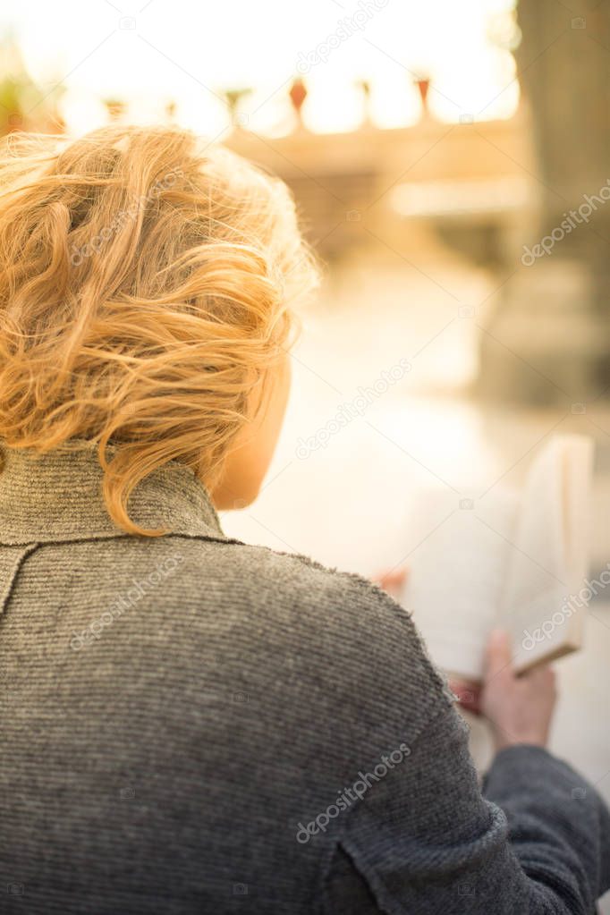 Stylish young woman in a gray coat outside the home, sitting on the sidewalk and reading the book. Her hair fluttering in the wind