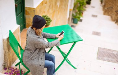 Yeşil sandalyede oturan ve sohbet için onun smartphone kullanarak ev dışında gri paltolu şık genç kadın