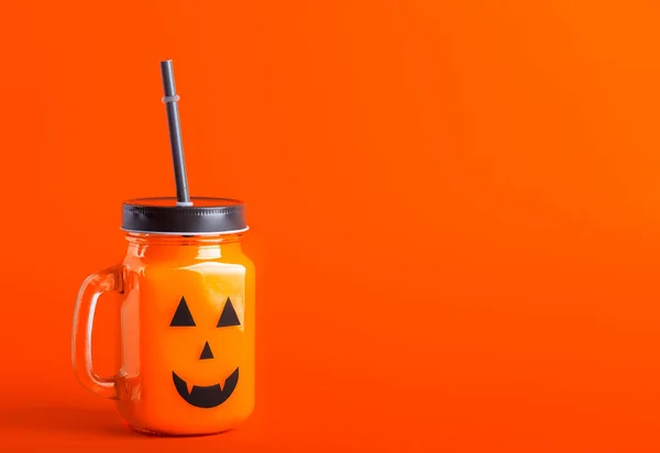 Halloween healthy pumpkin or carrot drink in the glass jar with scary face on a orange background