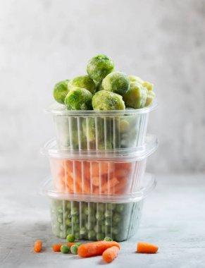 Frozen vegetables such as green peas, brussels sprouts and baby carrot in the storage boxes on the concrete gray background clipart