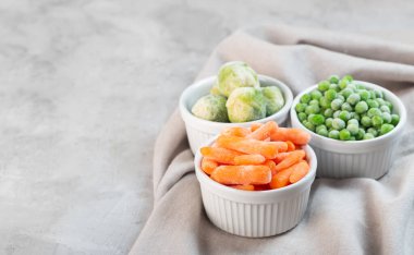 Frozen vegetables such as green peas, brussels sprouts and baby carrot in the white bowls on the concrete gray background with copy space clipart