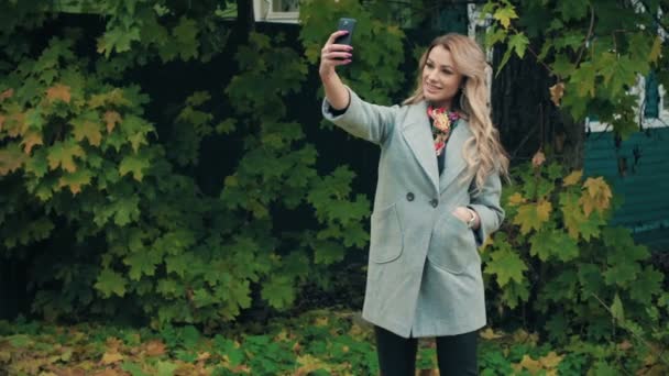 Blonde in gray coat doing selfies on smartphone in autumnal park — Stock Video