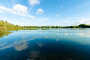 Durgun göl boyunca geniş bir manzara beyaz bulutlar ve mavi gökyüzü yüzeye yansıyor. Yoğun bir ağaç çizgisi ufku oluşturur..