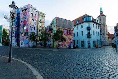 Şen şakrak şekilleri ve renkleri olan parlak bir bina sokak manzarasında göze çarpıyor. Çakıl taşları ve çevreleyen evler, olay yerini sabah ışığında tamamlıyor..