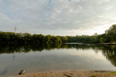 Yumuşak dalgalı sessiz bir göl, kıyı boyunca uzanan ağaçları yansıtır. Bulutlarla dolu bir gökyüzünün altında..