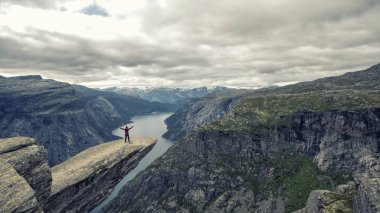 Kanyonda Ringedalsvatnet gölünün mavi su üzerinde ünlü Troltunga uçurumun kenarında cesur handstand yapma kırmızı ceketli genç adam dağcı. Norveç, yaz manzarası. Wanderlust kavramı.