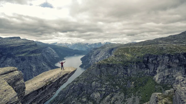 Young man climber in red jacket making intrepid handstand at the edge of famous Troltunga cliff over blue water of Ringedalsvatnet lake in canyon. Norway, summer scenery. Wanderlust concept.