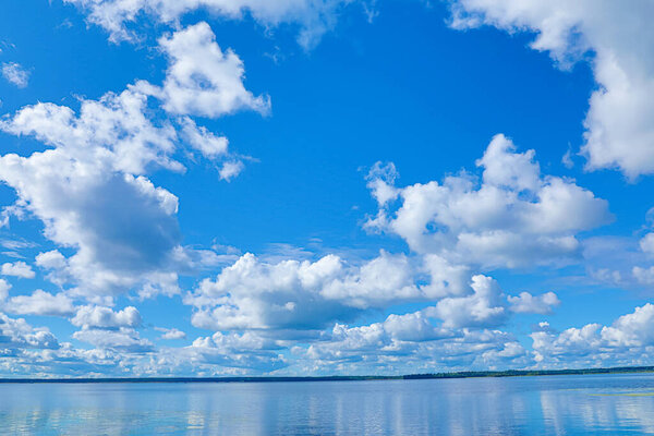 Clouds and blue sky above the water of lake, river or sea bay and far bank or shore with forest on the background in sunny summer or spring day